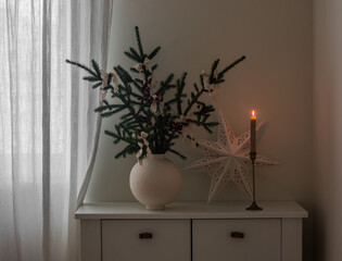 Christmas composition in a ceramic vase, paper star, burning candle on a white wooden chest of drawers in the living room