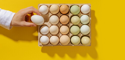 Aesthetic Easter Eggs in carton box on yellow, one in woman hand holding, minimal style banner with white beige green gradient colored Chicken egg, organic dyed eggs. Creative festive food concept