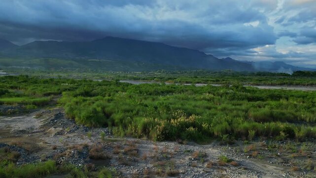 A forward ground aerial of grass fields near the Chico River reveals distant mountains shaded under clouds Bontoc, Nueva Vizcaya, Philippines