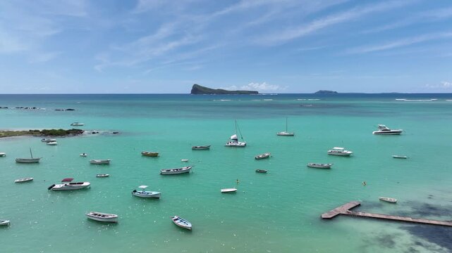 Drone shot in Mauritius, Cap Malheureux. The camera passes a tree in the foreground, revealing anchored yachts on an emerald ocean. Gunner&rsquo;s Quoin Island rising on the calm horizon in the background.