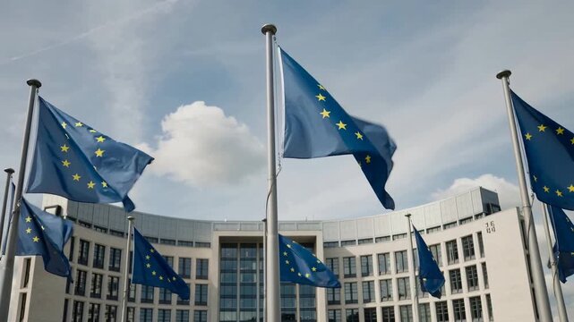 The image shows a row of European Union flags waving in front of a modern building. The building appears to be the headquarters of the European Commission.