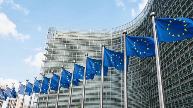 The image shows a row of European Union flags waving in front of a modern building. The building appears to be the headquarters of the European Commission.