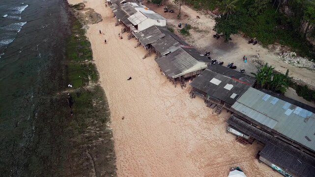 Cinematic drone view of tropical beach village in Indonesia, with wooden surf shacks along the coast, low tide reef exposed shallow water, moss patches and calm shoreline conditions in daylight.