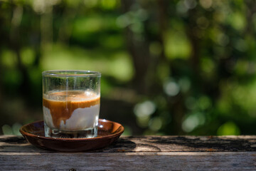 a small shot glass of chilled milk filled with hot espresso shot coffee floated over white creamy chilled milk put on wooden table in wild garden with sun shine and shade of green trees