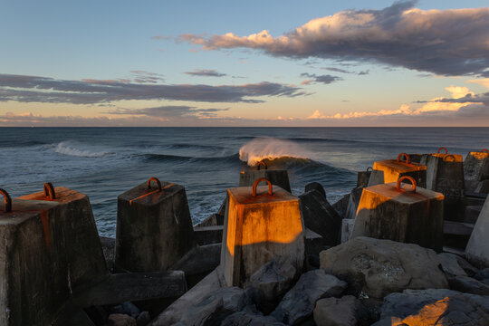 A breaking and sunlit wave with flying, windblown spray, catches the last rays of the sun next to a breakwater pier, with a blue sky and cloud backdrop at Ballina in New South Wales, Australia.