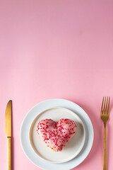 heart-shaped donut with pink icing and red sprinkles is presented on a white plate, flanked by a golden knife and fork, all centered on a solid pink background.