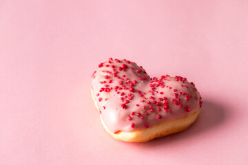 A close-up shot of a single heart-shaped donut with pink icing and red sprinkles, resting on a clean pink background, highlighting its texture and color.