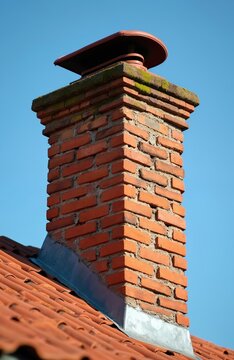 Red brick chimney with weathered top cap rises above orange tile roof against clear blue sky. Metal flashing joins chimney to rooftop. Old house structure detail.