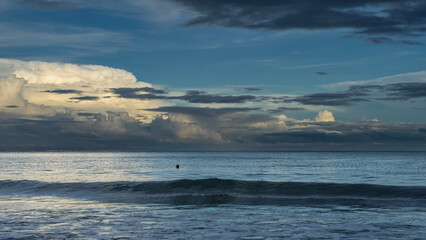 Obraz premium Early morning. The ocean waves are rolling towards the shore. The head of a floating man is visible in the sea. The clouds in the blue sky are illuminated with golden light. Malaysia. Borneo.