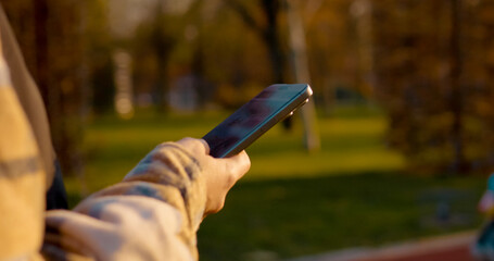 Leisurely using smartphone while walking in the park bench during golden autumn afternoon, woman browsing digital content in soft sunlight