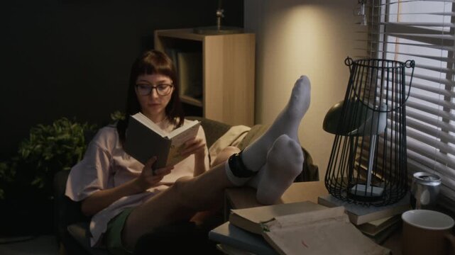 Young woman relaxing in cozy chair with her feet propped on table, wearing house arrest bracelet, reading book
