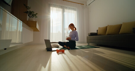 A redhead woman sits on a gym mat in her living room, engaging in an online fitness class with her laptop on a sunny morning while surrounded by dumbbells and her cat.