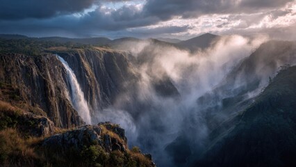 Majestic Waterfall Cascading Over Rocky Cliffs with Misty Clouds in Mountainous Landscape at Sunrise
