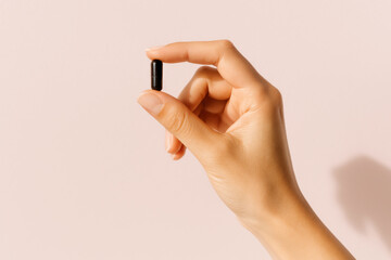 hand gripping a black capsule symbolizing medication vitamins or supplements for daily health wellness and medical care on a light pink background