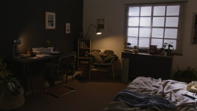 No people shot of interior of dimly lit messy bedroom with cluttered desk and armchair with blanket