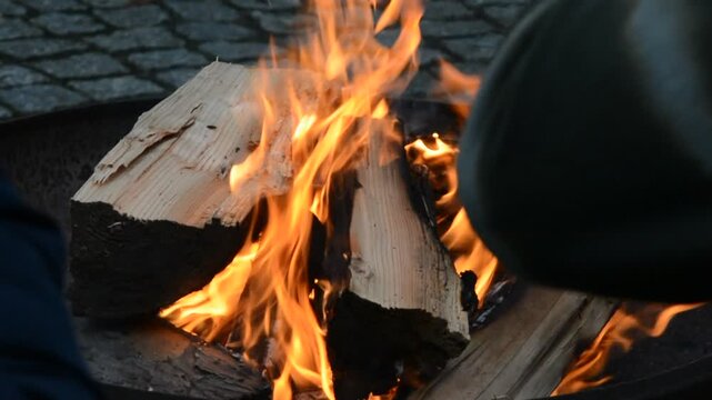Close-up of split wood logs burning with bright orange flames in a fire bowl against a blurred cobblestone background, creating a cozy warmth