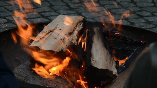 Close-up of split wood logs burning with bright orange flames in a fire bowl against a blurred cobblestone background, creating a cozy warmth