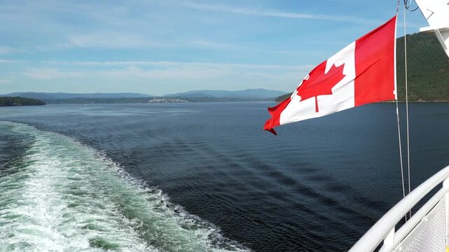Canadian flag waving in the wind on a ferry sailing from vancouver island to vancouver, british columbia, with the wake visible in the water and a sailboat in the distance