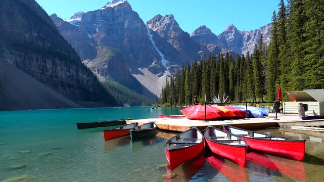 Majestic mountains surround the pristine turquoise waters of moraine lake, where vibrant red canoes await adventurers, nestled against a wooden dock under a clear blue sky