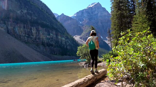 Young woman tourist walks carefully on a log over the turquoise water of the iconic moraine lake in banff national park, alberta, canada, enjoying the stunning view of the valley of the ten peaks