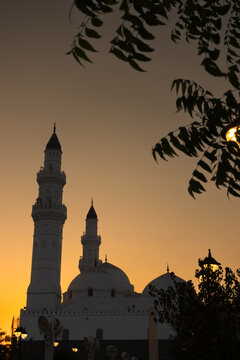 quba mosque with tall minarets and a dome silhouetted against a vibrant orange and yellow sunset sky, framed by dark tree branches.