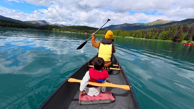 Mother wearing yellow beanie and life jacket paddling canoe with son on turquoise waters of maligne lake, surrounded by stunning jasper national park scenery in canadian rockies