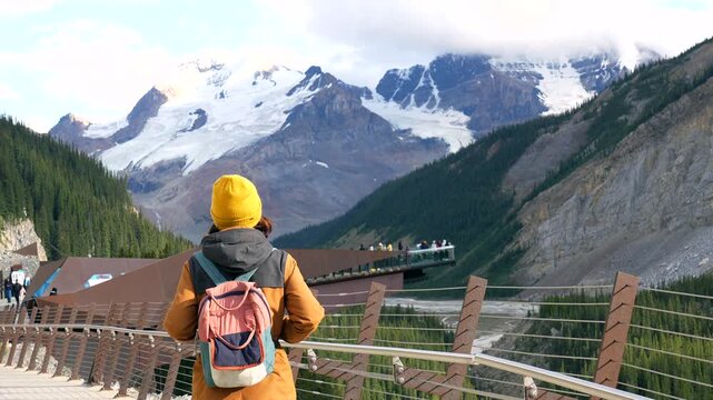 Hiker wearing yellow hat and backpack walking on skywalk at columbia icefield, viewing athabasca glacier and surrounding canadian rockies mountains in jasper national park, alberta