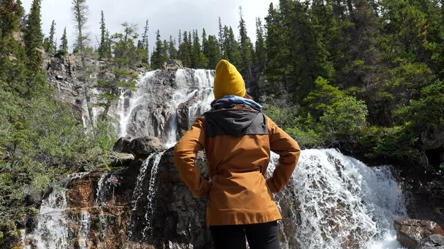 Female hiker wearing a yellow beanie and an orange jacket admires tangle creek waterfall on the icefields parkway in the canadian rockies, canada