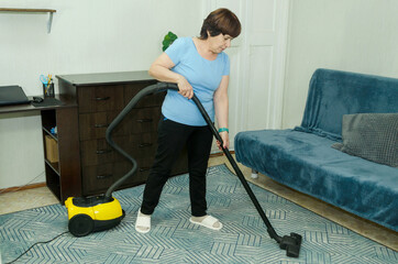 An elderly woman in red rubber gloves vacuums carpet near sofa and chest of drawers during daily house cleaning and dust removal