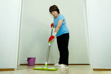 An elderly woman in protective gloves cleans floor with mop in room, symbolizing daily wet cleaning of the house.