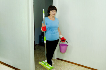 Elderly woman in red rubber gloves enters the room holding mop and bucket, ready to clean floors, illustrating usual house cleaning and home hygiene.