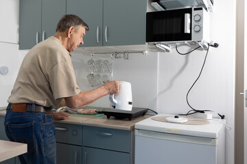 Senior man standing in modern kitchen pouring hot water from electric kettle into mug on table.