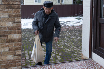 Elderly man walking with cane across brick courtyard, carrying shopping bag, winter setting