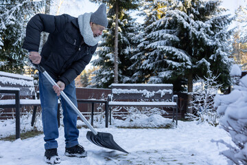 Elderly man clearing snow on outdoor path in winter park surrounded by trees benches cold seasonal environment.