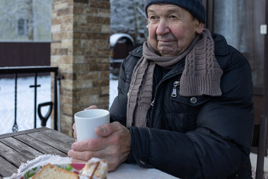 Elderly man drinking hot tea from mug while sitting outdoors at wooden table with simple meal during cold winter season.
