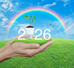Fototapeta premium 2026 white text with fresh tree leaves on soil and light bulb on green grass in hands over blue sky, white clouds and rainbow, Happy new year 2026 green ecology and saving energy concept