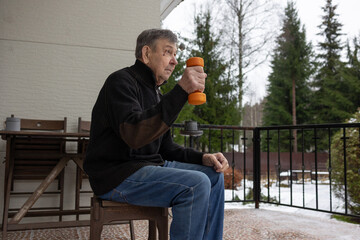 Elderly man sitting on chair holding orange dumbbell during simple home exercise on outdoor terrace with calm focused posture.