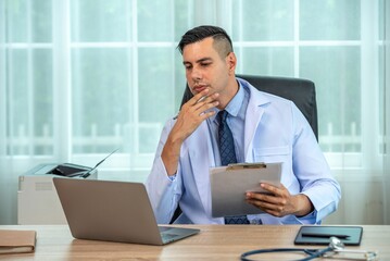 A male doctor in a white coat is sitting at his desk looking at a clipboard and thinking.