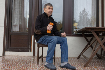 Elderly man sitting on chair holding orange dumbbell during simple home exercise on outdoor terrace with calm focused posture.