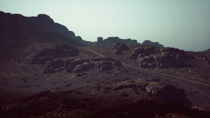 Obraz premium Boulders and rugged terrain create a striking scene as dusk settles over the mountains. A winding path leads through the rocky formations under a gentle mist, enhancing the atmosphere.