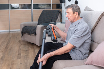 Senior man holding vacuum cleaner while resting on couch in cozy apartment interior showing everyday household activity.