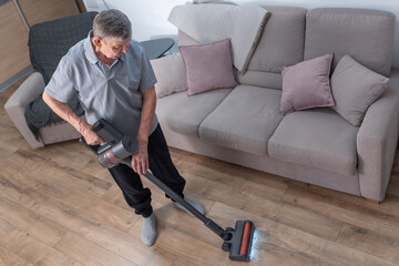 Cleaning elderly man vacuuming wooden floor in modern living room using cordless vacuum cleaner during everyday home routine
