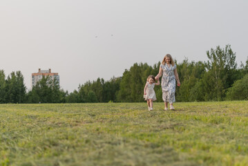 A young Caucasian woman and a little girl walk hand in hand through a grassy field. They are enjoying a sunny day outdoors, surrounded by trees and a distant building.