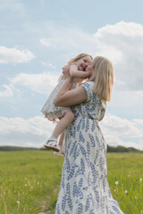 A young Caucasian woman with long blond hair holds a smiling girl in a white dress. They are in a green field under a blue sky with fluffy clouds.