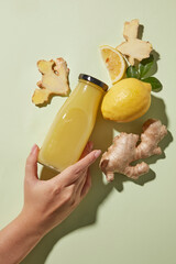 Flat lay with a hand holding a juice bottle among fresh ingredients - lemon and ginger roots. Ingredient of detox and healthy lifestyle storytelling, ginger shot product