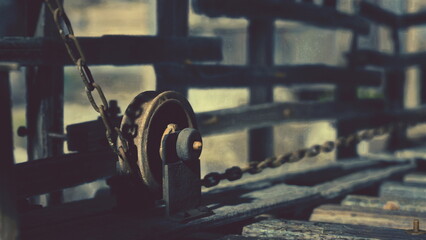 An old, rusty winch sits on weathered wooden beams at an abandoned construction site. The sunlight casts shadows, highlighting the aged metal and the surrounding neglected structures.