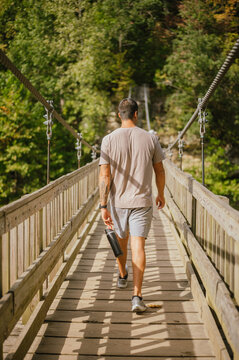 Backview of young man walking across suspension bridge holding water