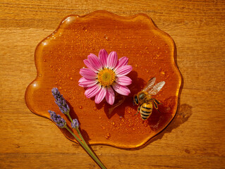 Overhead view of spilled golden honey puddle with a honey bee, pink cosmos flower, and lavender stalks on a rustic wooden table surface.