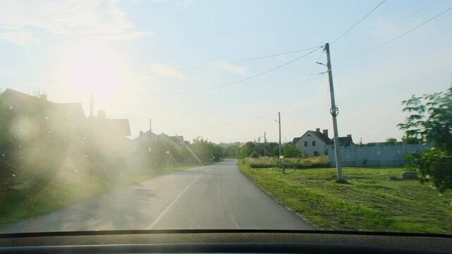 Narrow village road near low old houses through moving car window. Morning sun glares on glass and while neighborhood rolls past slowly