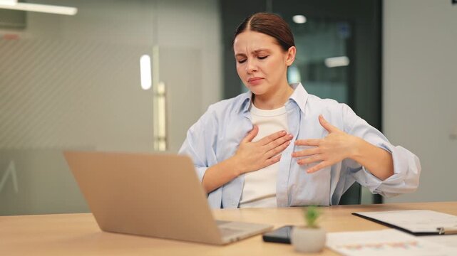 Woman working on laptop experiences sudden, intense chest pain. Distress and worry evident, indicating potential health concern or anxiety attack during office hours.